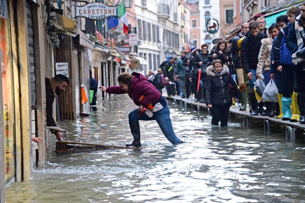 A woman tries to cross a flooded street as people walk on a trestle bridge during high water, in Venice, northern Italy, 15 November 2019. Photo: Andrea Merola / ANSA / AP