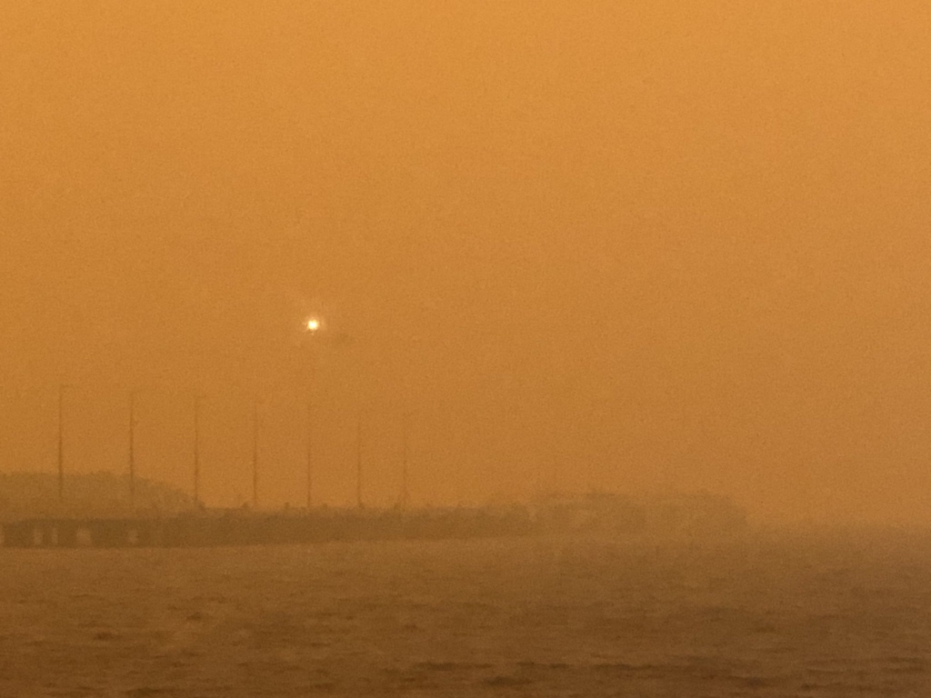 Barely visible through the smoke, a helicopter circles above the wharf in the town Eden, 4 January 2020. The entire town was ordrered to be evacuated as bushfires closed in. Photo: Brett Mason / SBSNews