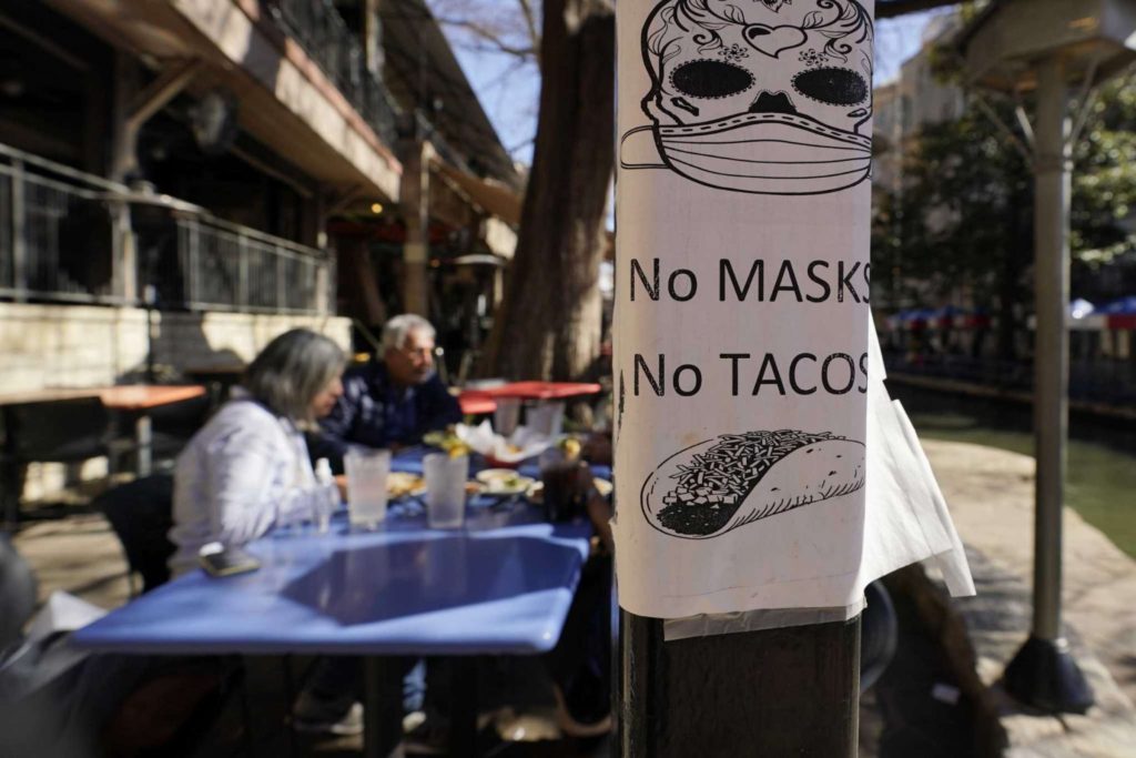 A sign requiring masks is seen near diners eating at a restaurant on the River Walk, in San Antonio, Texas, on 3 March 2021. The sign shows a Dia de los Muertos skull wearing a mask, with the words, “No masks, no tacos”. Texas Gov. Greg Abbott lifted the statewide mask mandate and business capacity limits without consulting his medical experts. Photo: Eric Gay / AP