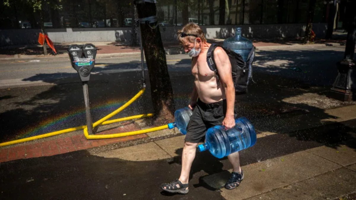 A man carrying multiple jugs of water walks through a sprinkler during a period of record-breaking temperatures in Vancouver on Monday, 28 June 2021. Photo: Ben Nelms / CBC