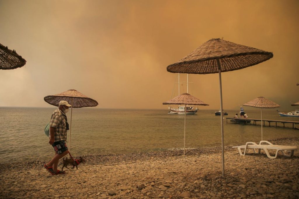 A man walks with his dog on the beach of smoke-engulfed Mazi area as wildfires rolled down the hill toward the seashore, in Bodrum, Mugla, Turkey, Sunday, 1 August 2021. More than 100 wildfires have been brought under control in Turkey, according to officials. The forestry minister tweeted that five fires are continuing in the tourist destinations of Antalya and Mugla. Photo: Emre Tazegul / AP Photo