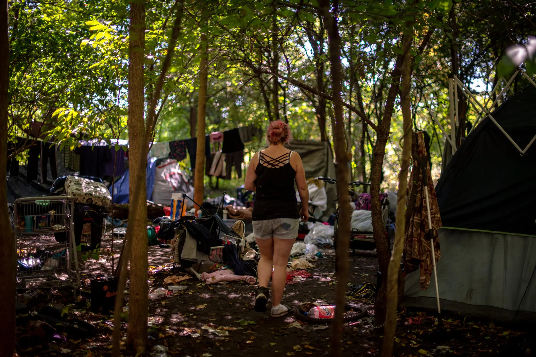 A view of a camp inhabited by people living with drug addiction. Belongings are strewn about the forest floor and a tent is set up at right. a woman, her back to the camera, limps along among the belongings, wearing a spaghetti-strap shirt, shorts, and her hair is dyed silvery lavender. Photo: Hilary Swift / The New York Times